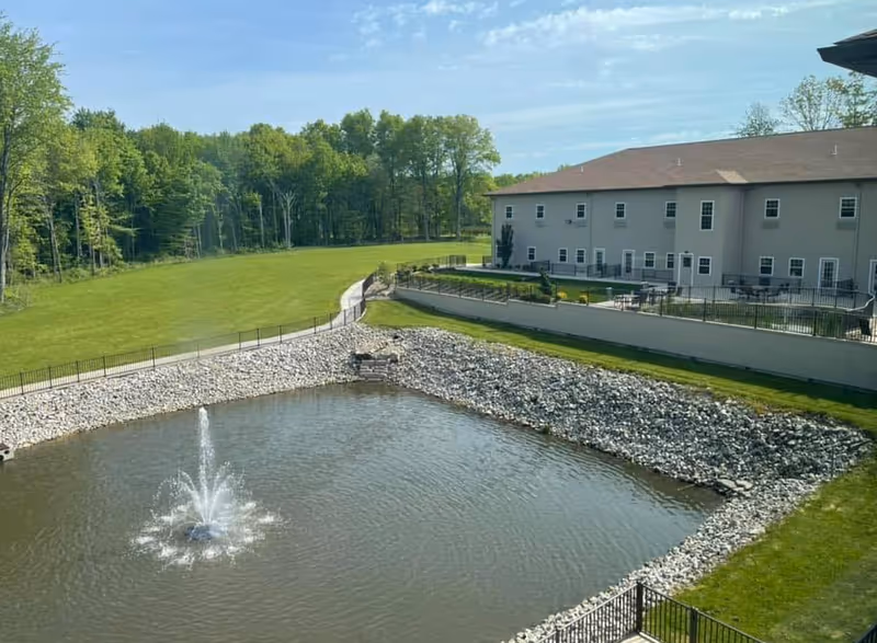 A landscaped pond with a central fountain in front of a two-story building, bordered by rocky shores, lawns, a walkway and trees.