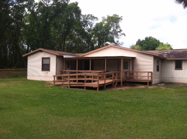 Single-story beige building with a wooden ramped porch and a grassy yard, set against a backdrop of trees.
