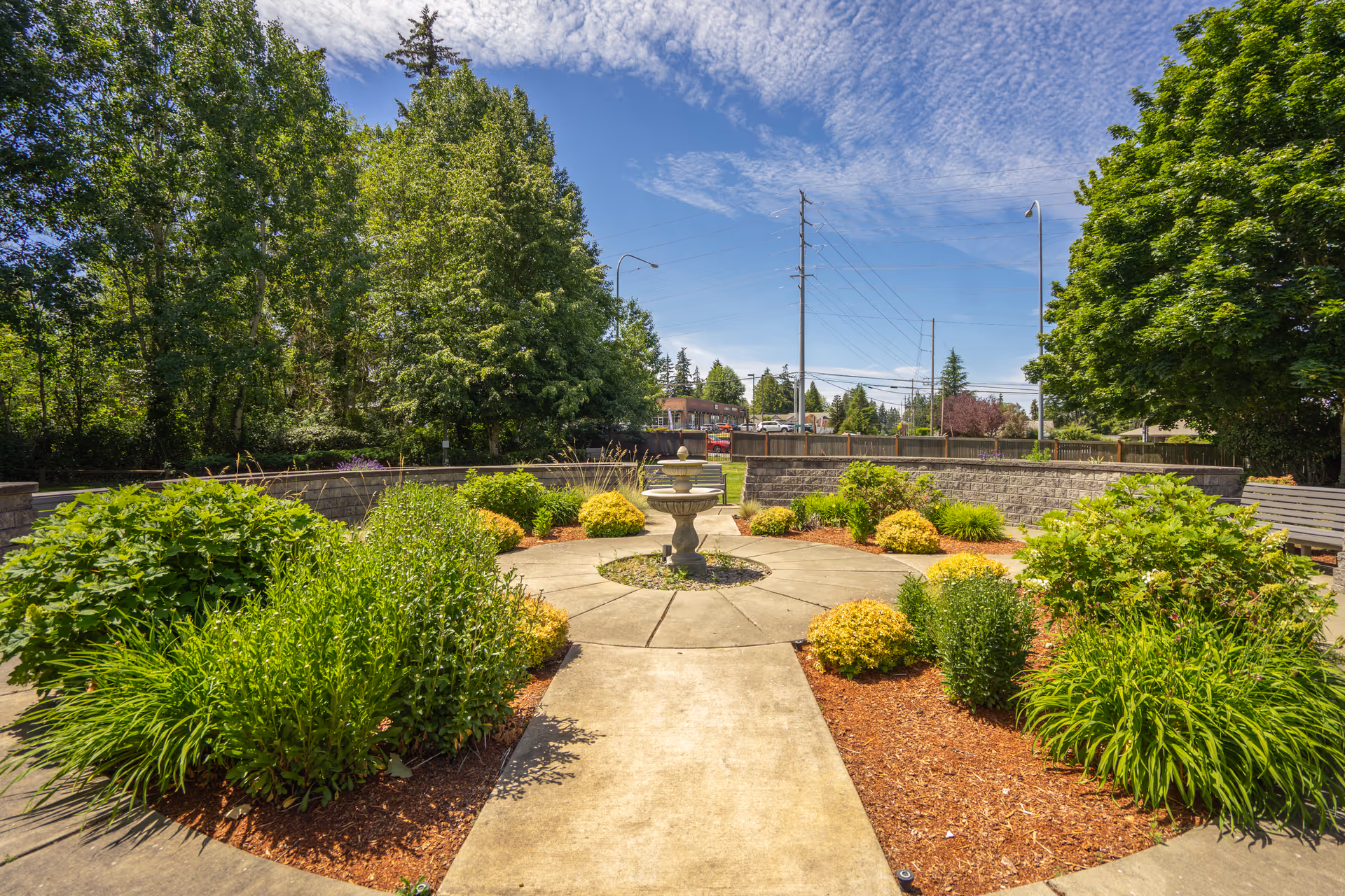 A landscaped outdoor garden area with a circular concrete pathway surrounding a central stone fountain. The garden is filled with various green shrubs and plants, bordered by mulch. There are tall trees and a stone wall enclosing the garden. The sky is partly cloudy with blue patches visible.