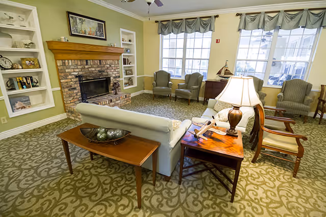 A cozy communal living room with sofas and armchairs arranged around a brick fireplace and side tables with a lamp.