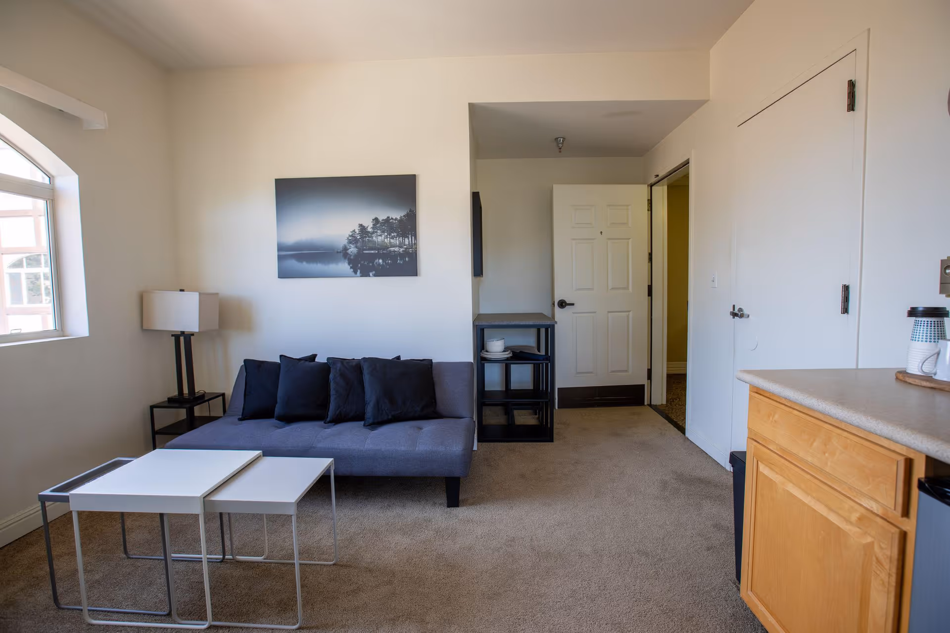 A small living area in an assisted living facility featuring a gray sofa with four dark cushions, a floor lamp with a white shade on a black side table, two nested white coffee tables, a black shelving unit with dishes, a closed white door, and a wooden kitchen counter with a coffee maker and mug. A window with an arched top lets in natural light, and a black and white landscape photograph hangs on the wall above the sofa.