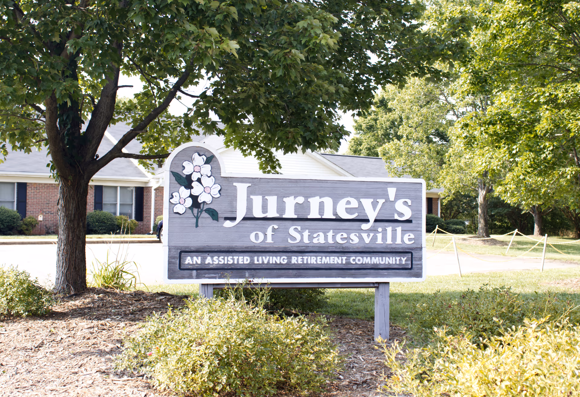 Outdoor view of a wooden sign for Jurney's of Statesville, an assisted living retirement community, surrounded by greenery and trees with a brick building in the background.
