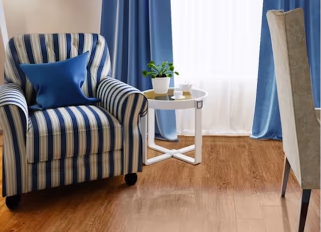 A cozy corner of a living room featuring a blue and white striped armchair with a blue cushion, a small white round side table with a potted plant and a cup, light wooden flooring, blue curtains, and a beige upholstered dining chair partially visible.