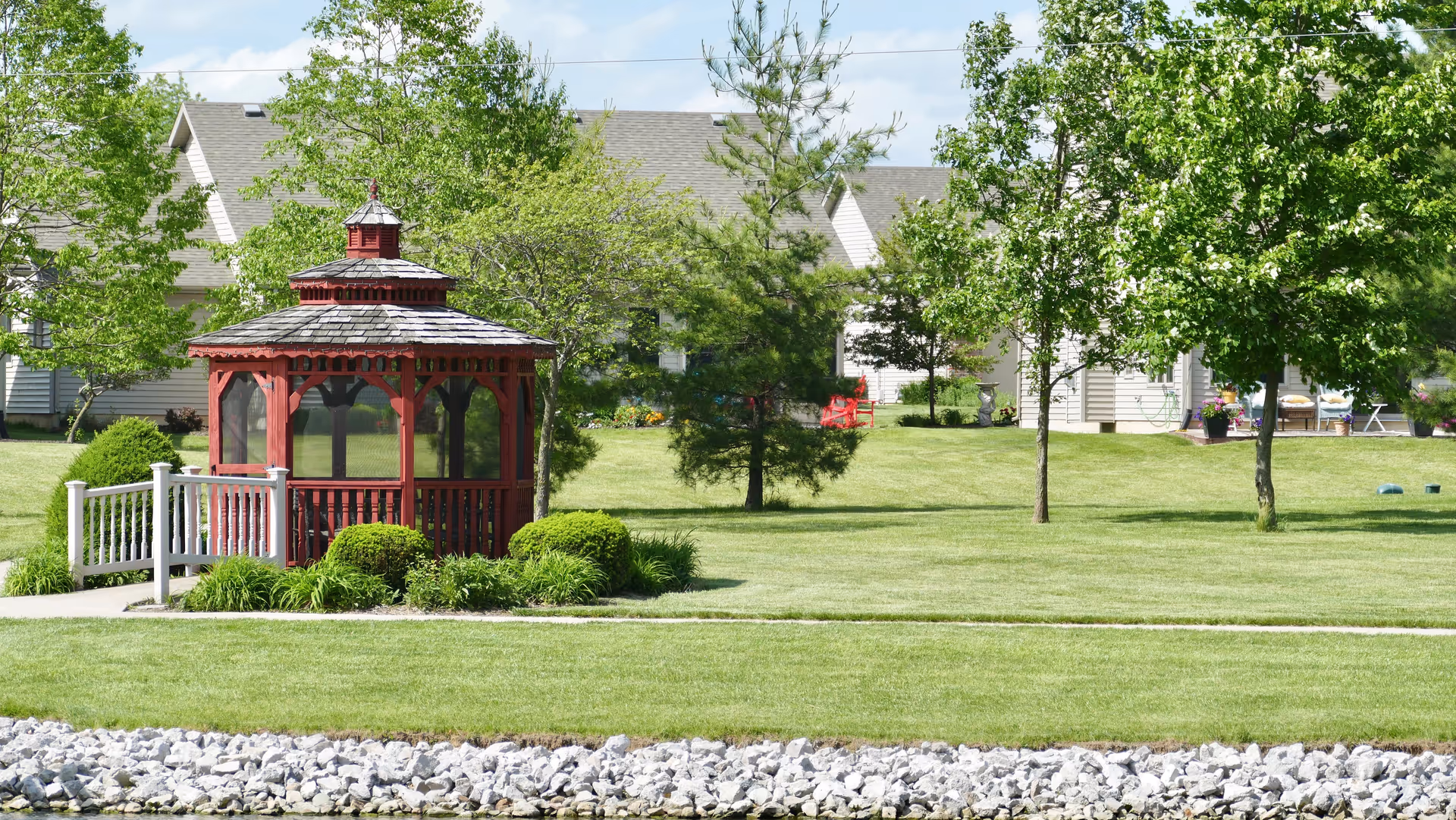 A well-maintained outdoor garden area featuring a red wooden gazebo with a shingled roof, surrounded by green grass, bushes, and trees. In the background, there are light-colored residential buildings with pitched roofs and some outdoor furniture visible.