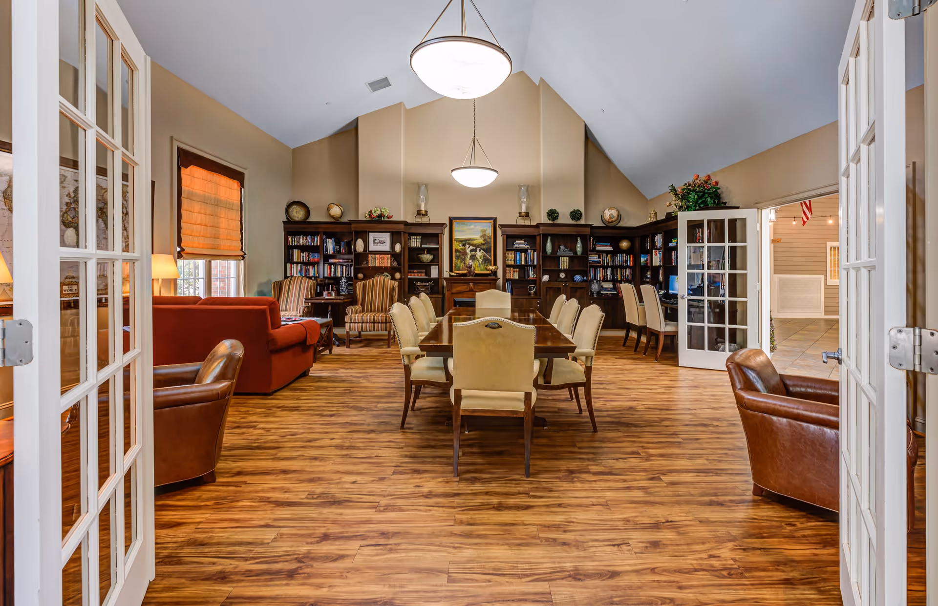 A spacious, well-lit common area with wooden flooring, featuring a large wooden table surrounded by beige upholstered chairs in the center. The room has bookshelves filled with books and decorative items along the back wall, two striped armchairs, a red sofa, and brown leather chairs. French doors open into the room, and there are two hanging ceiling lights providing illumination.