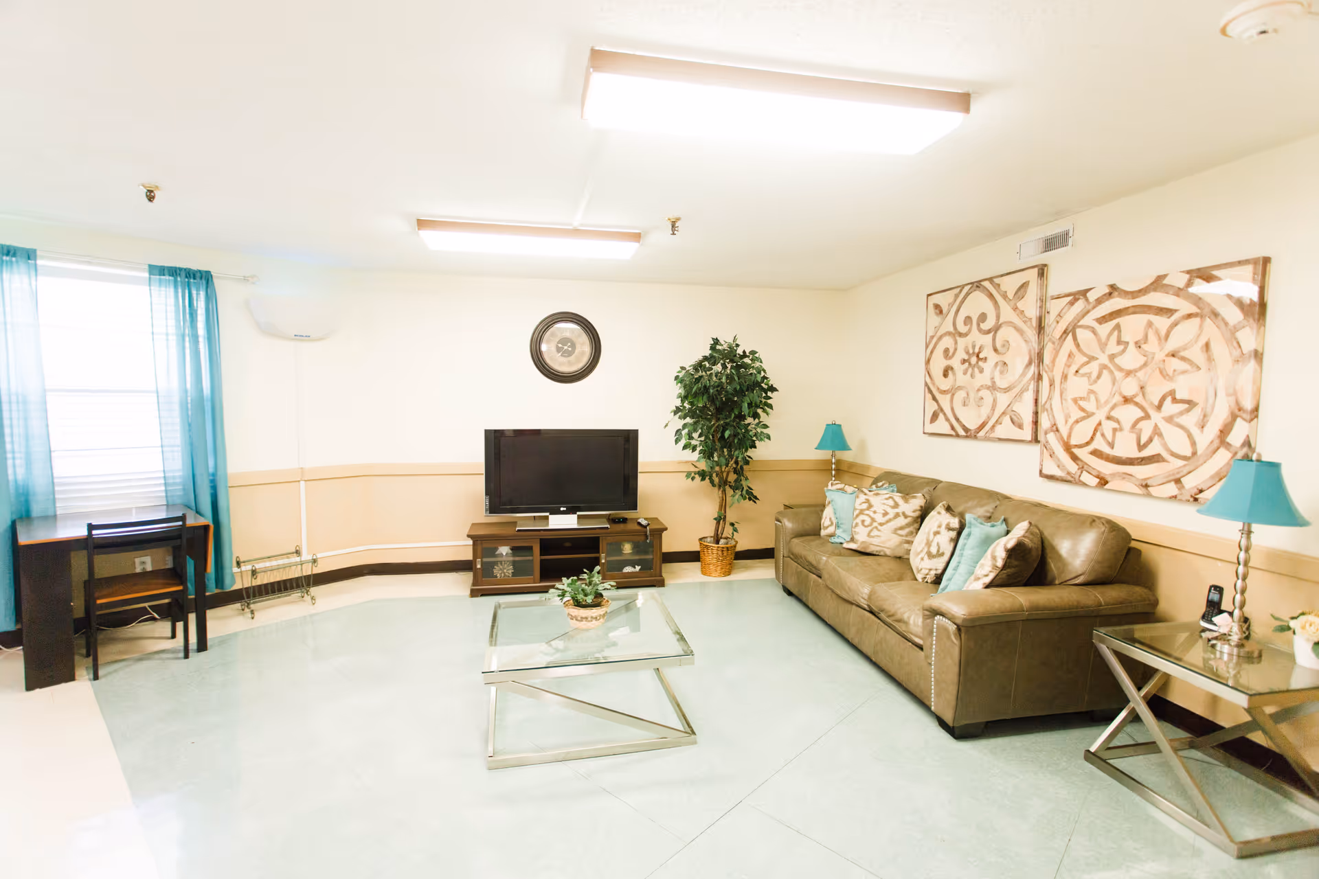 A bright living room with a brown leather couch adorned with decorative pillows, two teal lamps on glass side tables, a glass coffee table with a small plant, a flat-screen TV on a wooden stand, a potted plant in the corner, a small desk with a chair near a window with teal curtains, and two large framed decorative wall art pieces.