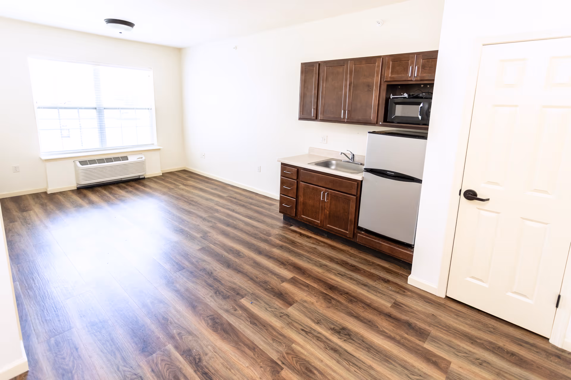 A bright, empty room with wood-style flooring, a large window with blinds, and a small kitchenette featuring dark wood cabinets, a sink, a microwave, and a mini refrigerator. A white door is visible on the right side of the image.