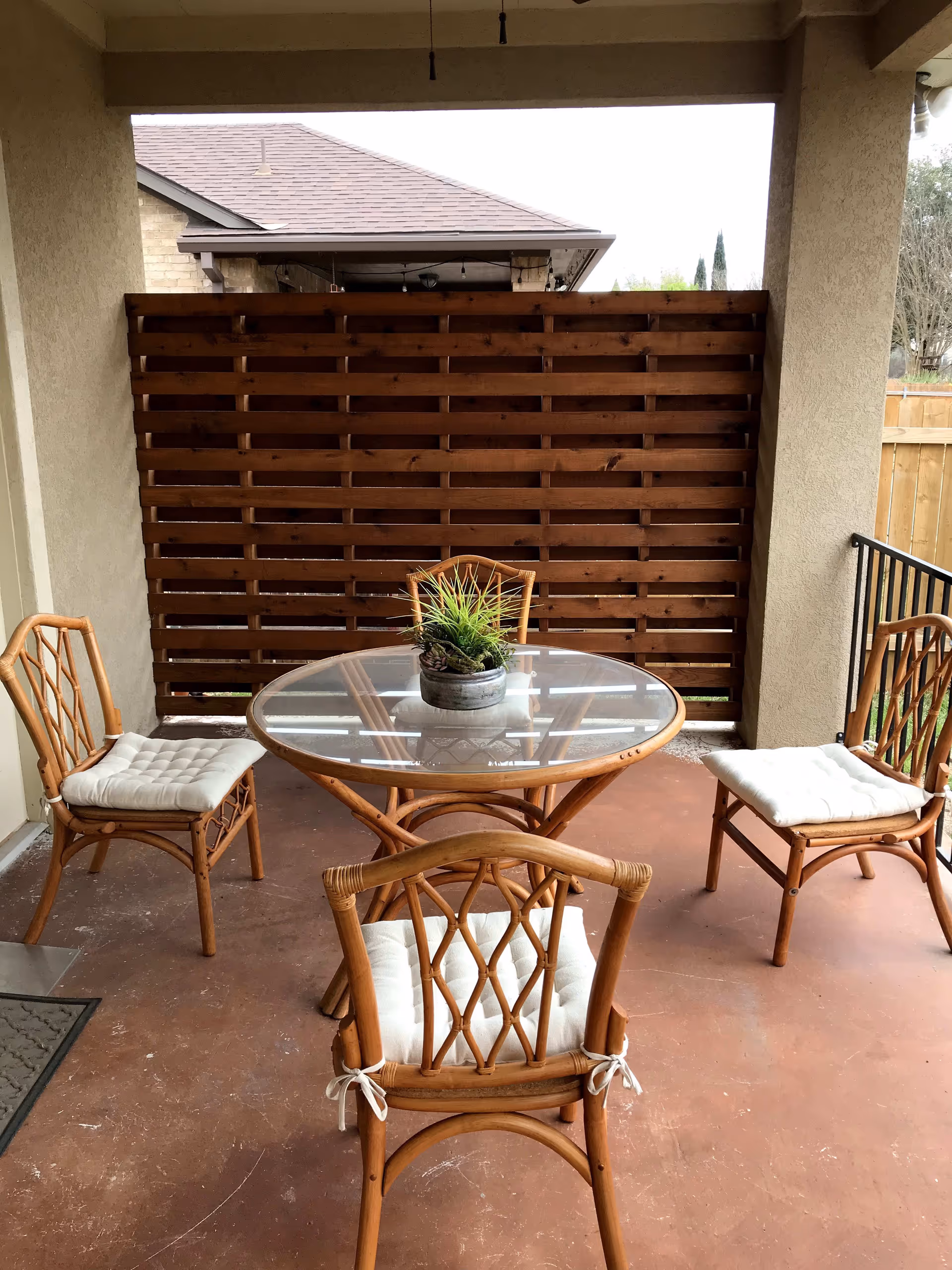 Covered outdoor patio area with a round glass-top table and four wooden chairs with white cushions. A small potted plant is placed in the center of the table. The patio has a reddish-brown floor and a wooden privacy screen on one side, with a view of a neighboring house roof and some trees in the background.