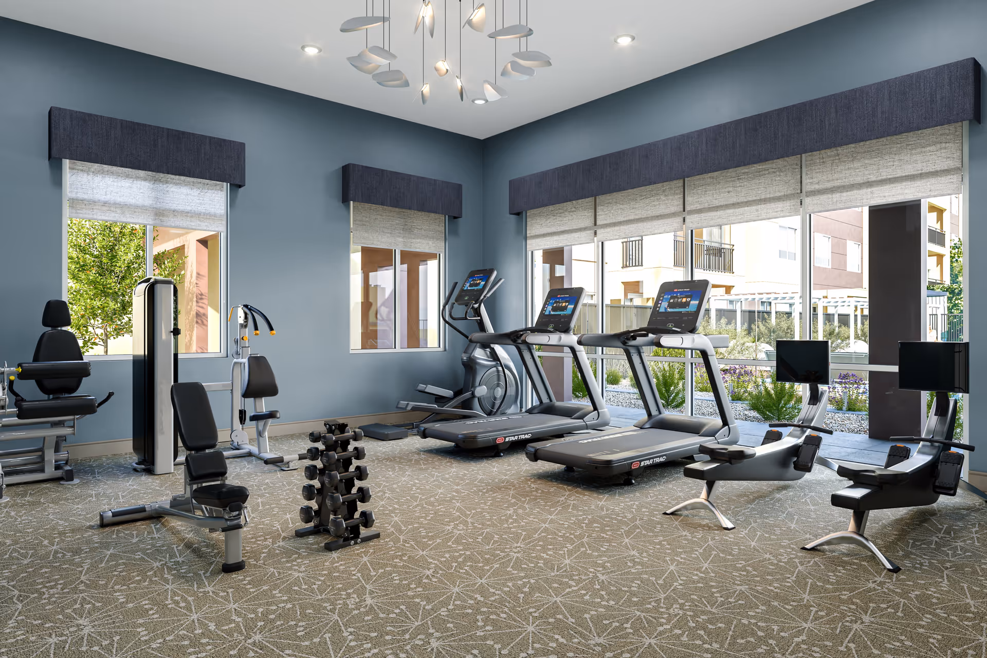 A fitness room with exercise equipment including two treadmills, two rowing machines with screens, a weight machine, a bench, and a rack of dumbbells. The room has large windows with views of greenery and buildings outside, blue walls, and a modern ceiling light fixture.