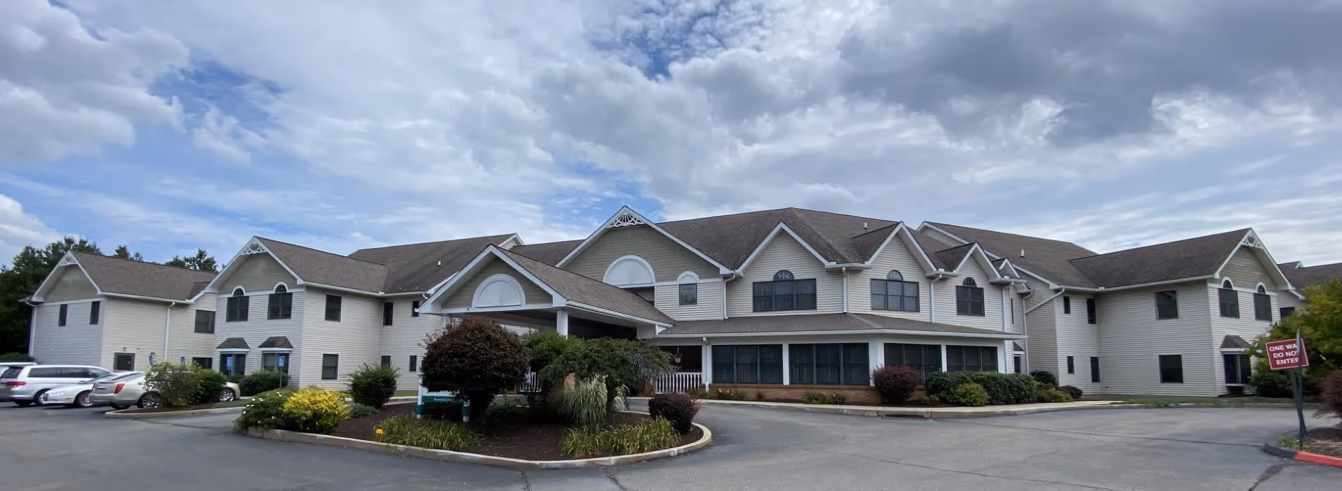 Exterior view of a large two-story senior living facility building with beige siding and multiple gabled roofs under a partly cloudy sky. The building is surrounded by a paved driveway and landscaped areas with bushes and small trees. Several parked cars are visible on the left side.