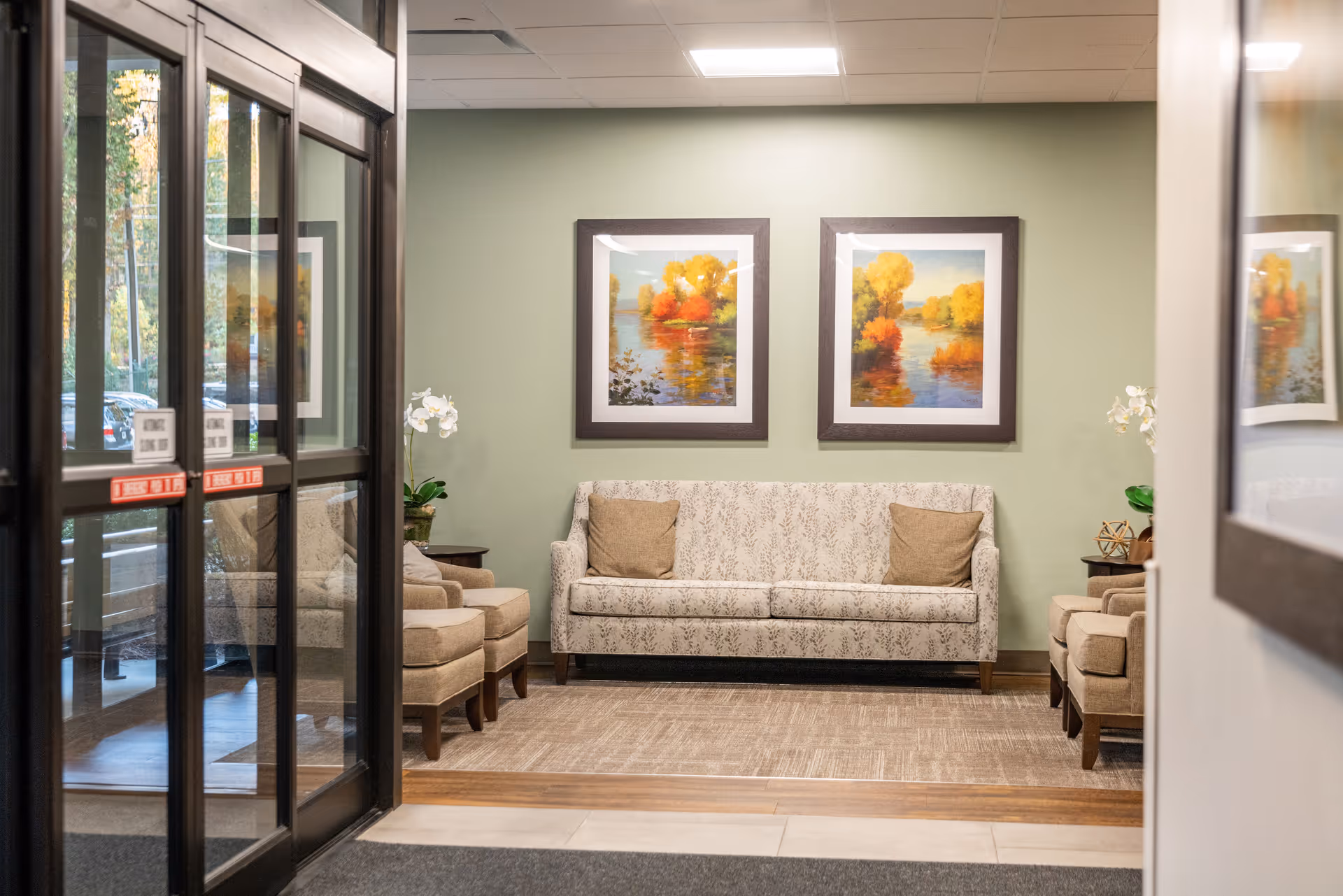 A cozy seating area in a senior living facility with a patterned sofa and two armchairs, each with a brown cushion. The walls are painted light green and decorated with two framed paintings of autumn trees by a lake. There are side tables with plants and decorative items on either side of the sofa. The area is viewed through glass doors with reflections of the outdoor parking lot.