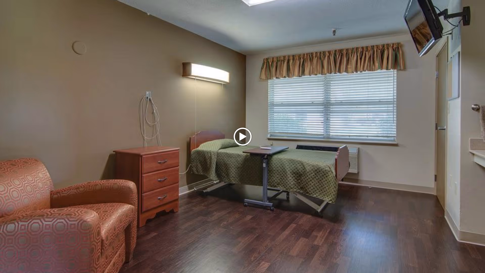 A senior living facility bedroom with a single bed covered in a green quilt, a wooden nightstand with three drawers, a patterned armchair, a wall-mounted light fixture, a window with blinds and a valance, a wall-mounted television, and a small sink area.