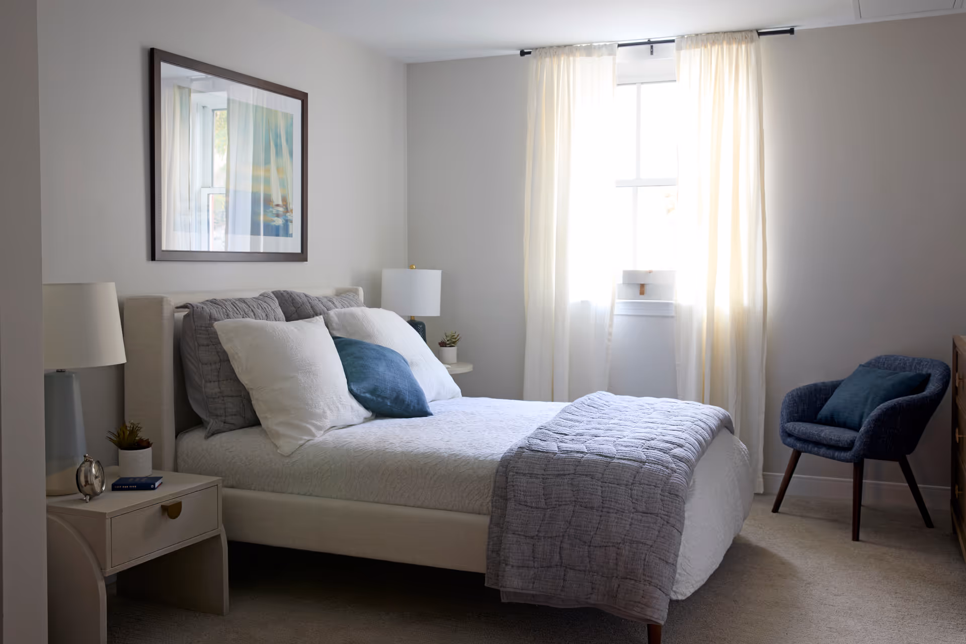 A neatly made bedroom with a bed, bedside tables and lamps, a window with light curtains, and a blue accent chair.