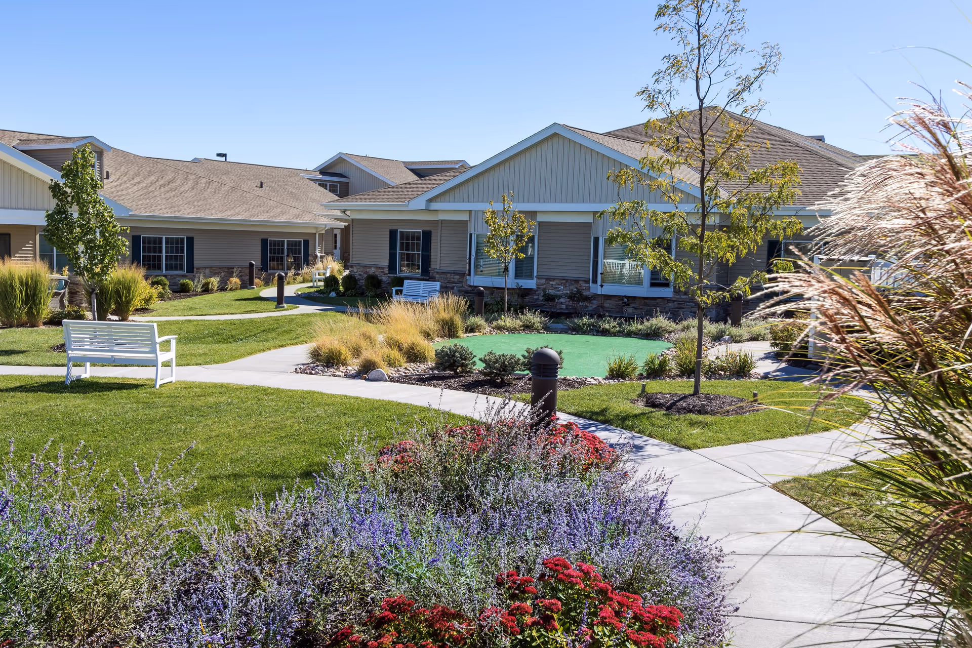 A landscaped outdoor garden area at CountryHouse Residence featuring green lawns, colorful flower beds with purple and red flowers, small trees, and paved walking paths. Beige residential buildings with stone accents and multiple windows surround the garden. White benches are placed along the paths for seating under a clear blue sky.