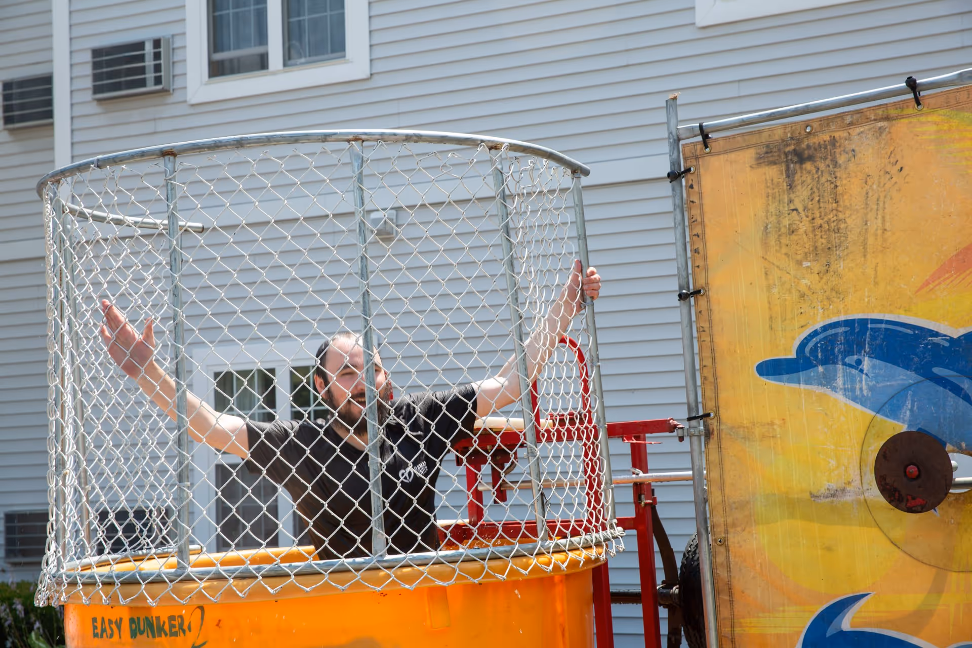 Man inside a chain-link dunk tank outdoors beside a building.