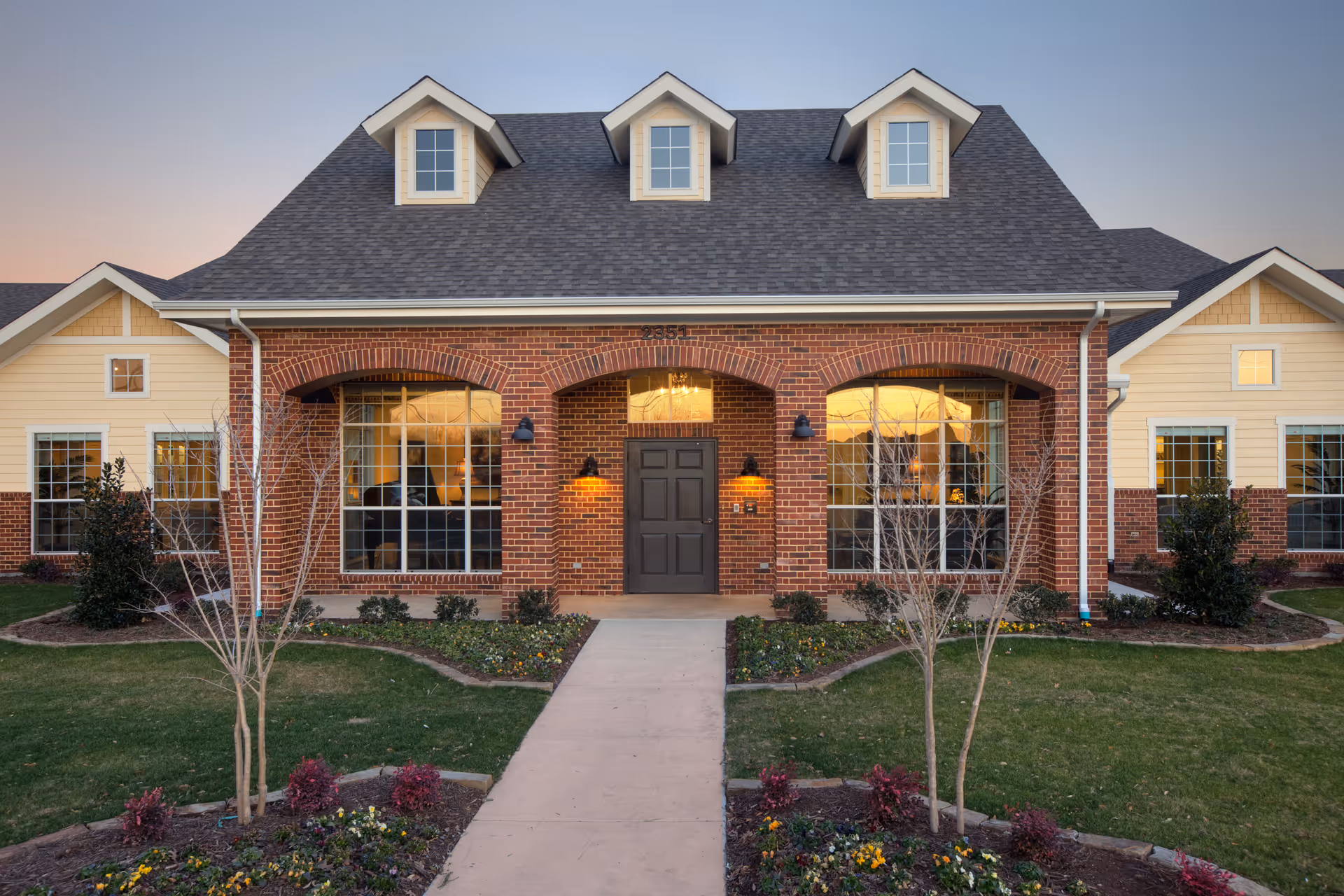 Brick building with three dormer windows and central front entrance, surrounded by landscaping