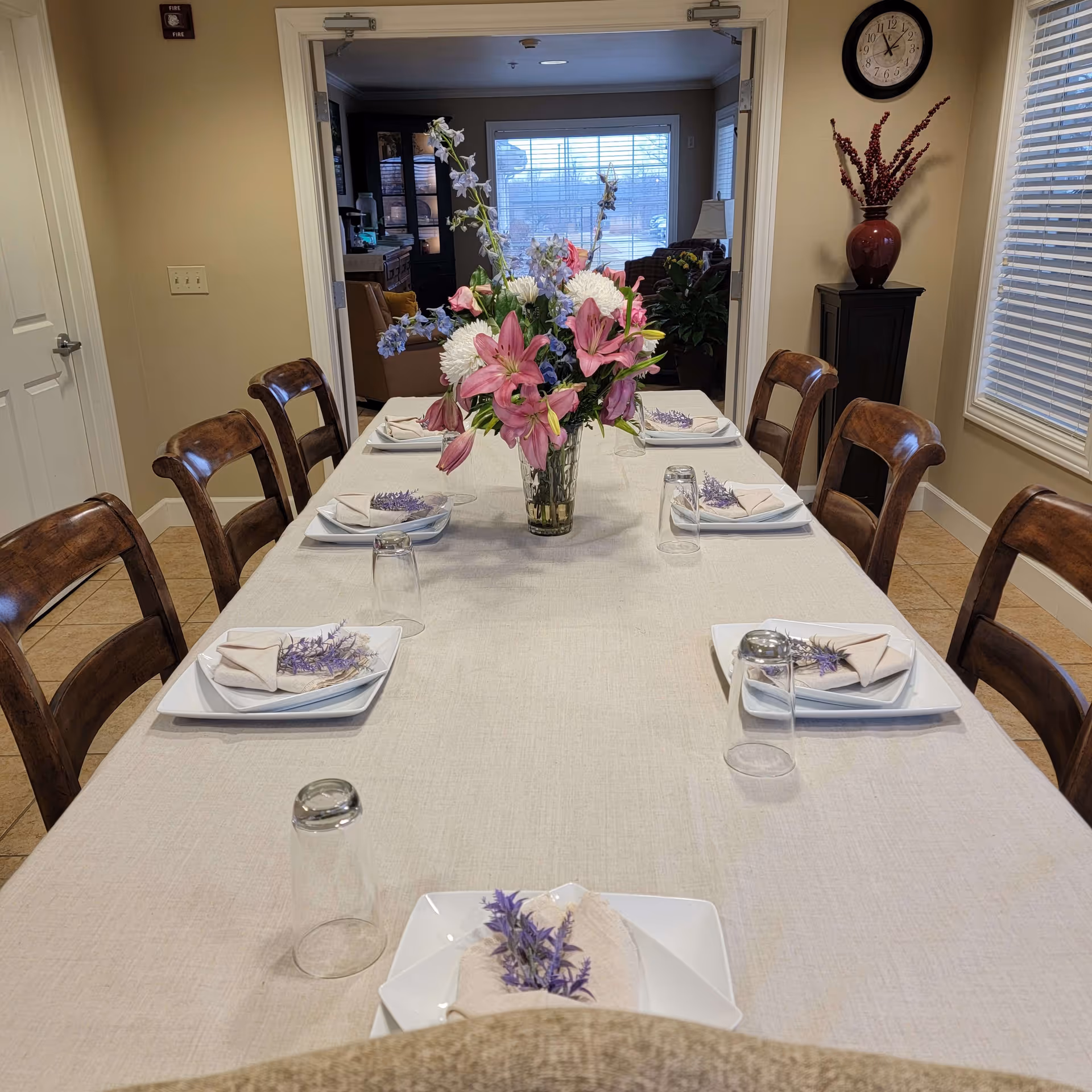 A dining room table set for six with white plates, beige napkins decorated with purple flowers, and upside-down drinking glasses. A large floral centerpiece with pink lilies and white flowers is in the middle of the table. The room has wooden chairs, beige walls, a clock on the wall, a window with blinds, and a vase with red branches on a small cabinet.