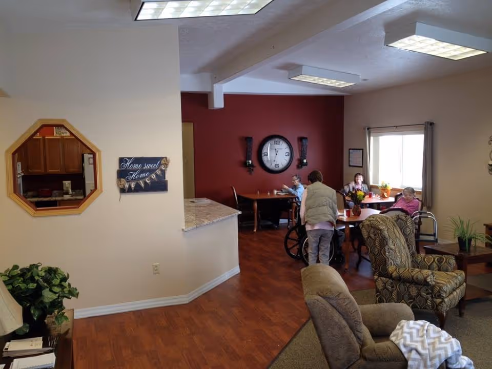 Interior common room with lounge chairs, tables and a few residents gathered by a red accent wall with a large clock.
