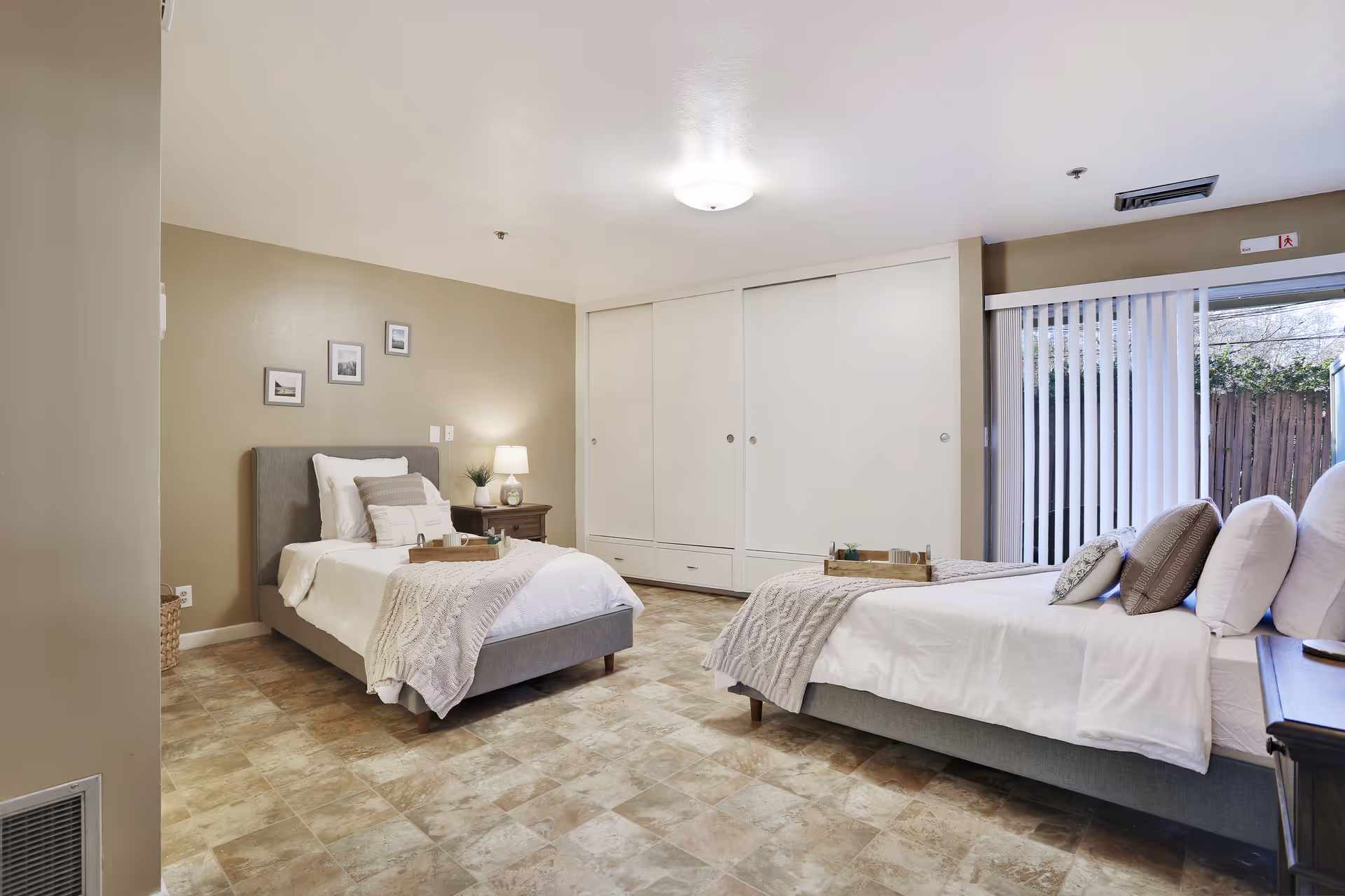 A bright bedroom with two single beds, each with white bedding, gray knitted throws, and decorative pillows. There is a nightstand with a lamp and a small plant between the beds. The room has beige walls, a tiled floor, three framed pictures on the wall, and a large closet with white sliding doors. Vertical blinds cover a sliding glass door that leads outside to a fenced yard.