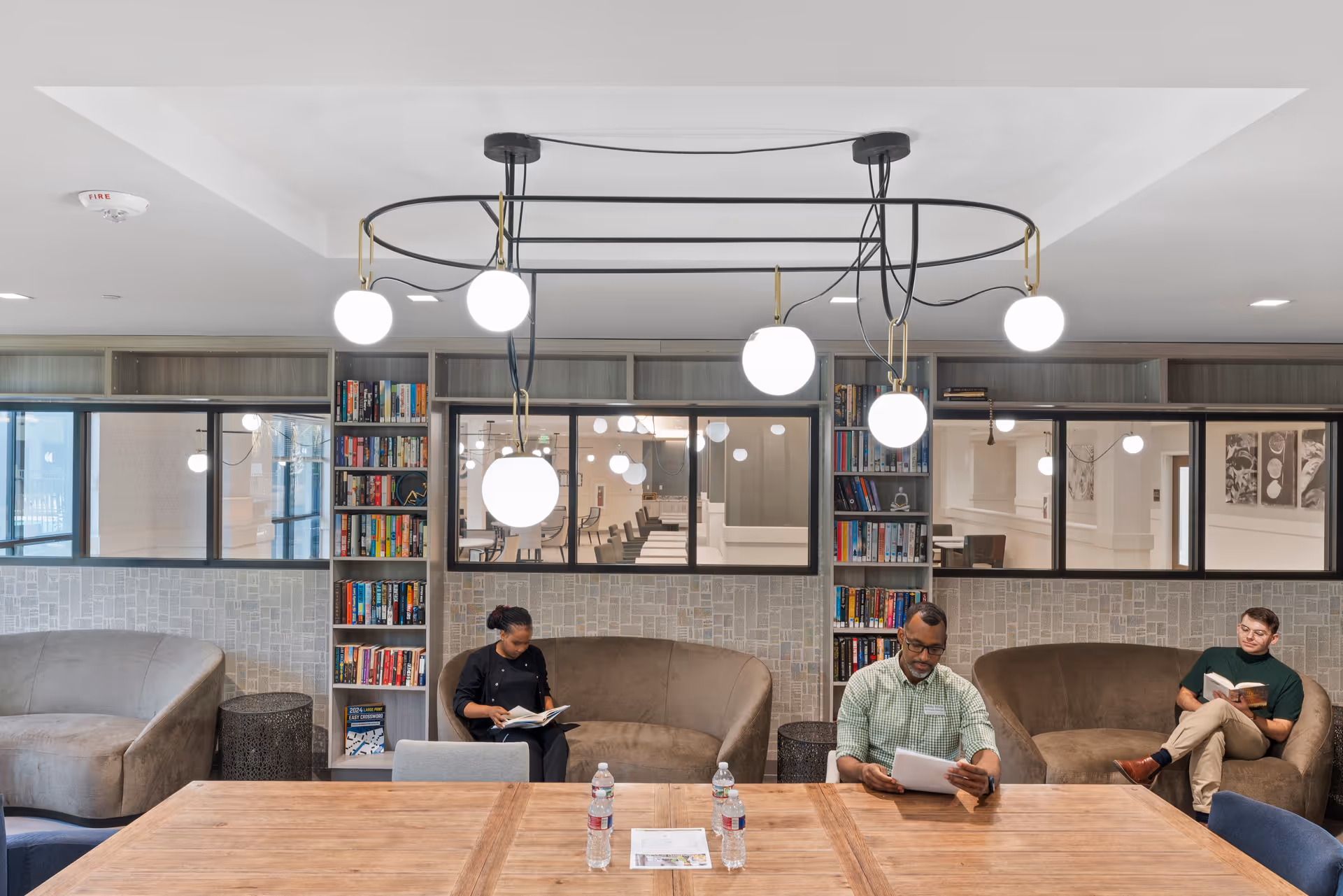 A modern common area with three people sitting on curved sofas reading books. The room features built-in bookshelves filled with books, large windows looking into another room, and a contemporary chandelier with round white lights hanging from the ceiling. A wooden table with water bottles and papers is in the foreground.