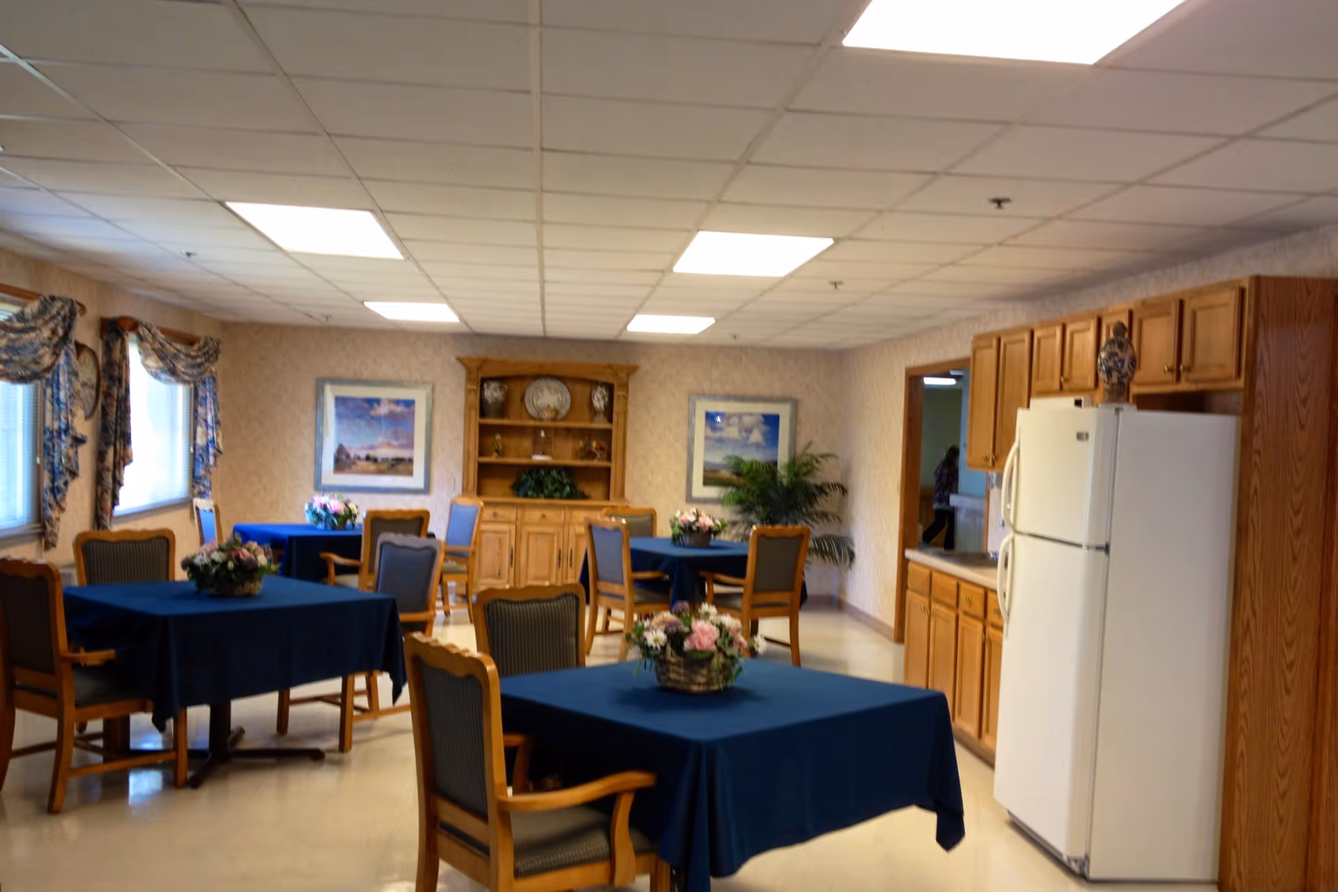 Dining room with tables covered in blue tablecloths, wooden chairs and floral centerpieces, and a kitchenette with cabinets and a refrigerator.