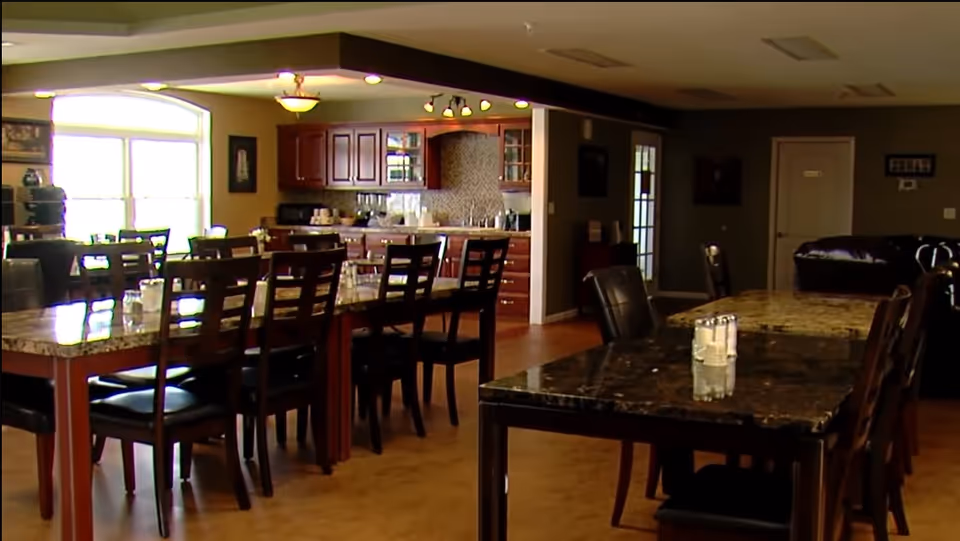 Interior view of a dining area with multiple tables and chairs. The room features a kitchen area with wooden cabinets and a tiled backsplash in the background. The tables have a marble-like surface and are set with salt and pepper shakers. There is a large window letting in natural light and a seating area with a dark couch in the back.