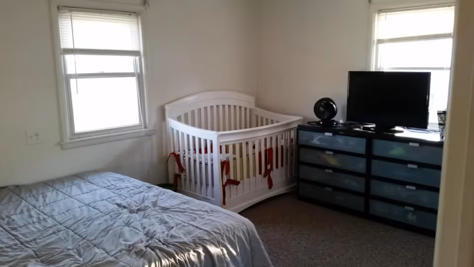 Small bedroom with a bed, a white baby crib, and a dresser topped by a TV between two windows.