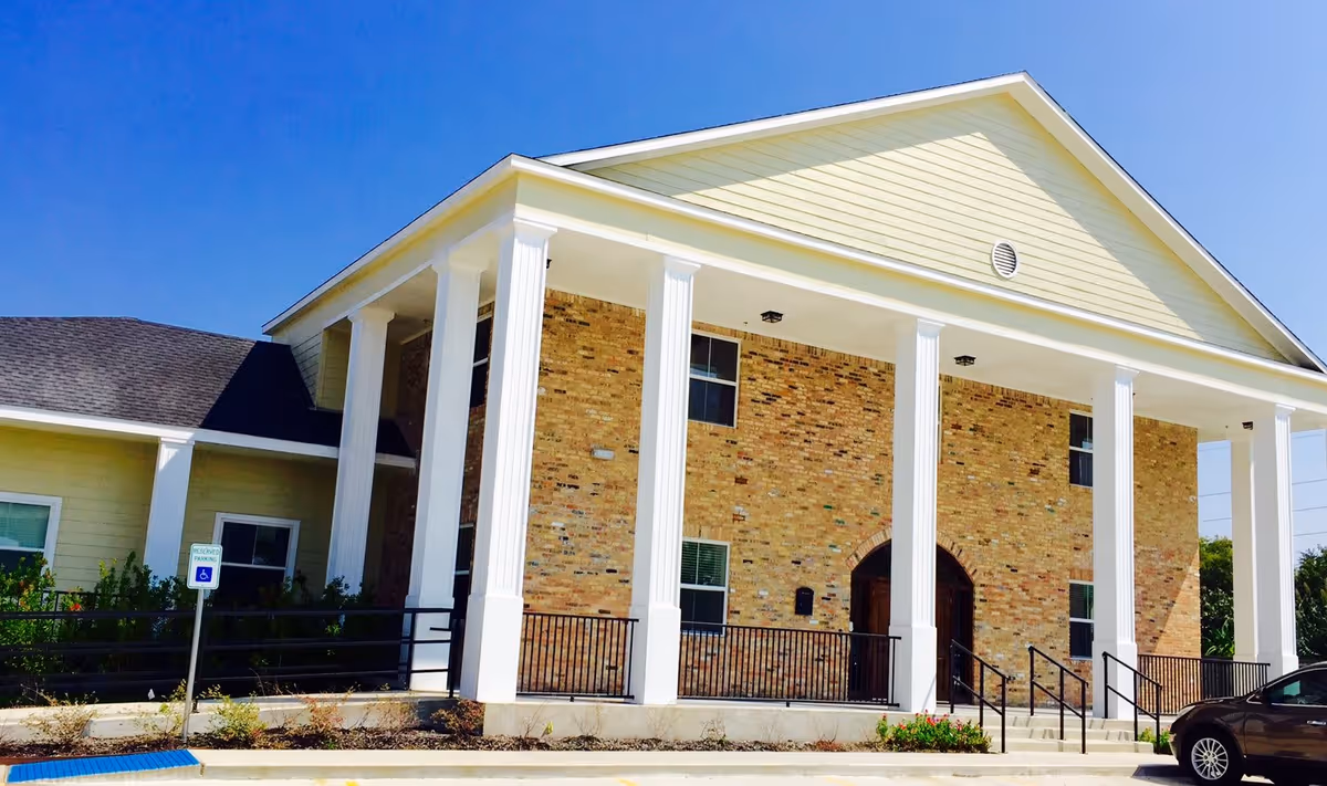 Exterior view of a senior living facility building with a brick facade, large white columns supporting a covered entrance, and a ramp with railings. There is a handicapped parking sign and a car parked on the right side under a clear blue sky.