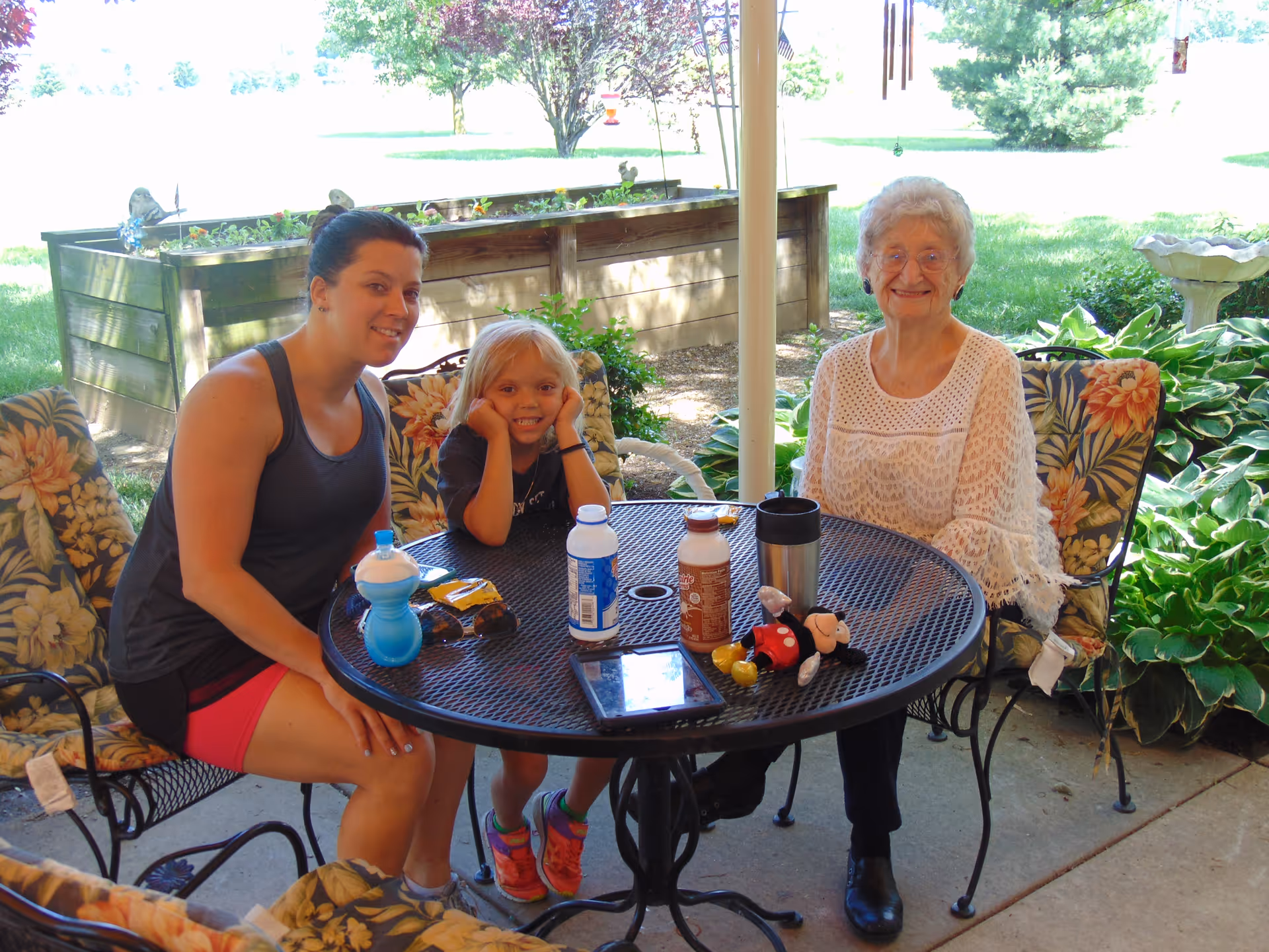 Three people sitting around a round outdoor metal table with floral cushioned chairs. An elderly woman, a young woman, and a young girl are smiling at the camera. The table has various items including drinks, snacks, a tablet, and a Mickey Mouse toy. The setting is a shaded patio with greenery and garden beds in the background.