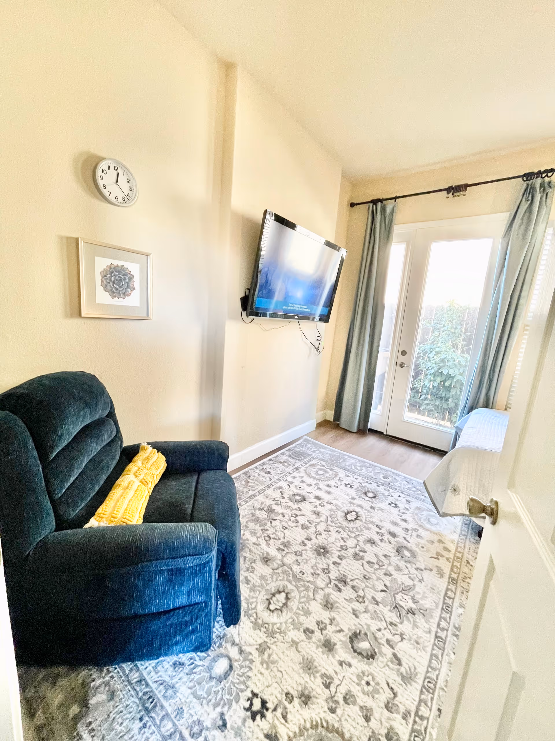 A small living room with a dark blue armchair with a yellow blanket on it, a wall-mounted flat screen TV, a patterned area rug, a wall clock, framed artwork, and glass doors with light blue curtains leading outside.