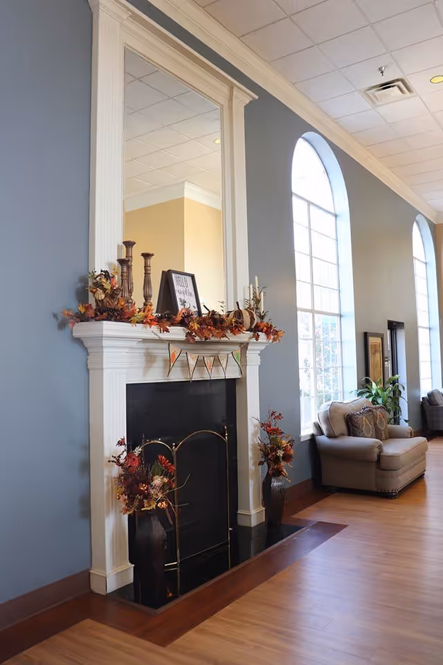 A cozy interior space featuring a white fireplace decorated with autumn-themed garlands, candles, and a framed sign. The fireplace is flanked by two vases with fall floral arrangements. To the right, there is a large arched window letting in natural light and a comfortable beige armchair with patterned cushions. The walls are painted blue and the floor is wooden.