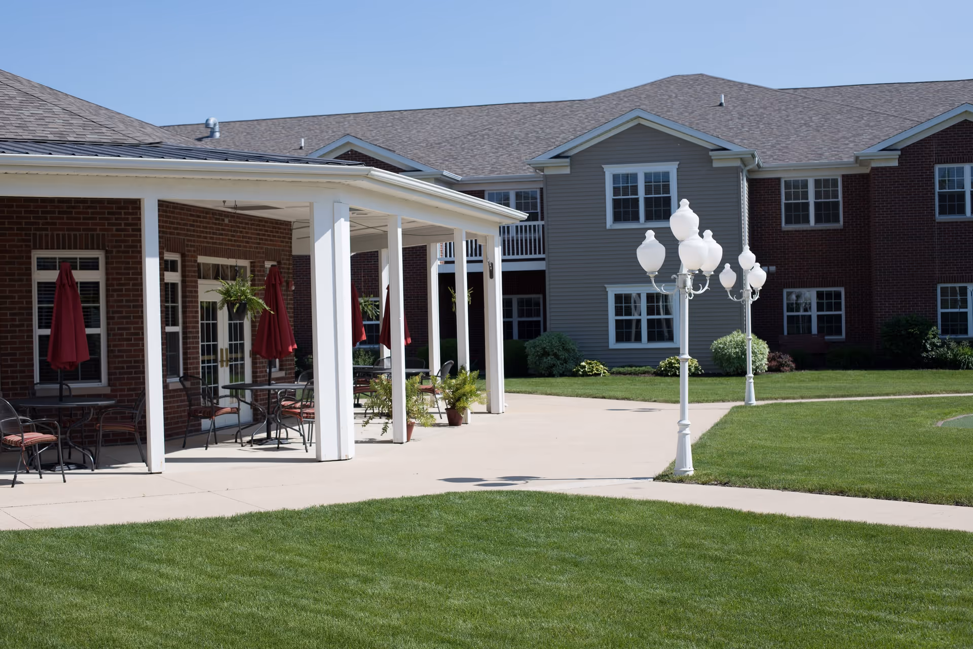 Outdoor patio area with tables, chairs, and red umbrellas next to a brick and siding building under a clear blue sky. There are white lamp posts on a concrete walkway surrounded by well-maintained green grass.