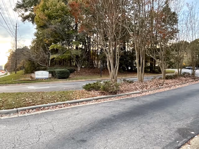 Driveway entrance with landscaped trees, fallen leaves, and a small stone sign for Brookdale Cary.
