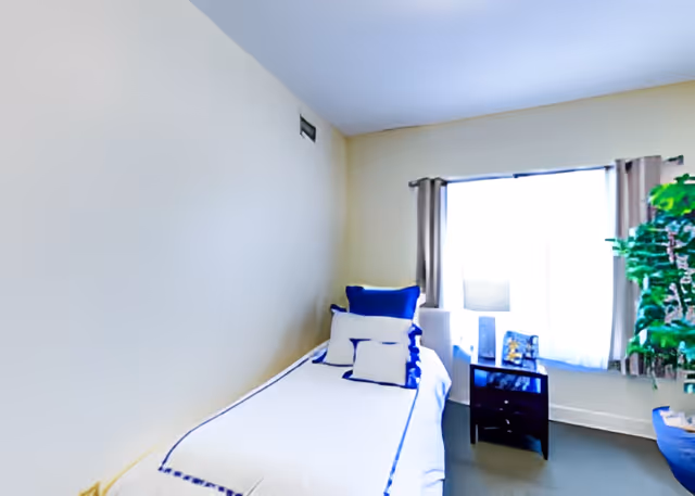 A small bedroom with a single bed against a beige wall. The bed is made with white bedding accented with blue trim and several pillows in white and blue. Next to the bed is a small dark wooden nightstand with a lamp and a few decorative items. A large window with beige curtains allows natural light to fill the room. There is a green potted plant near the window adding a touch of nature to the space.