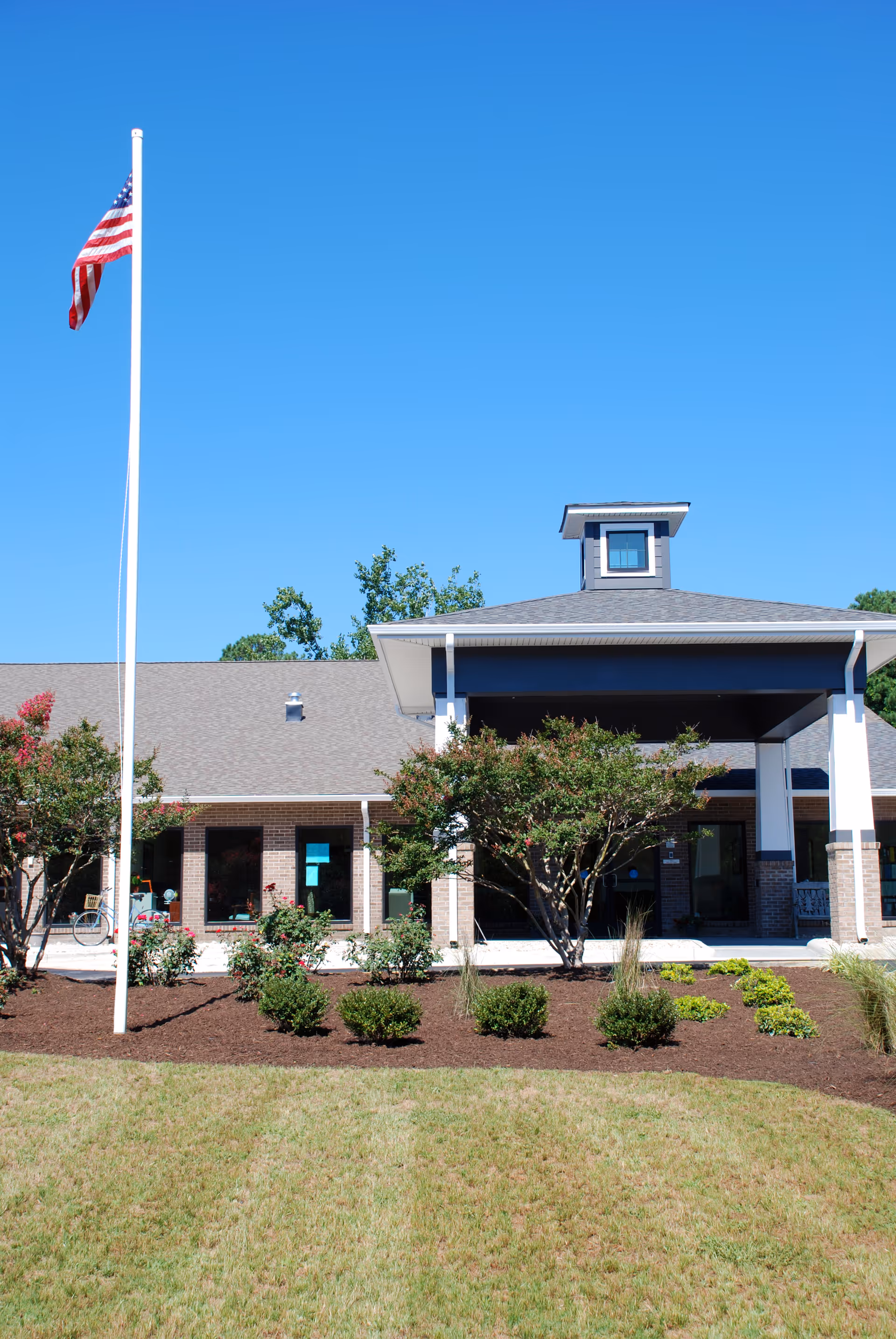Front entrance of a single-story brick senior living building with landscaping and an American flag on a tall flagpole under a clear blue sky.