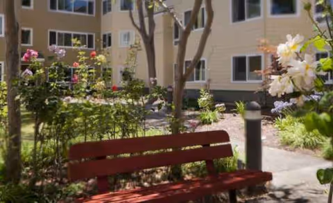 Red bench in a sunny courtyard with flowers and trees near a multi-story building.