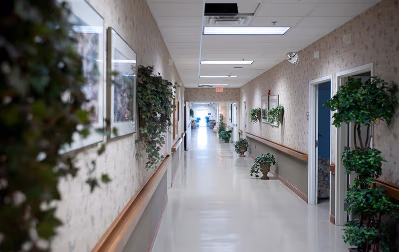 A long, clean hallway in a senior living facility with beige walls decorated with framed pictures and green plants. The hallway has handrails on both sides and several open doorways leading to rooms. The floor is shiny and light-colored, and ceiling lights illuminate the corridor.