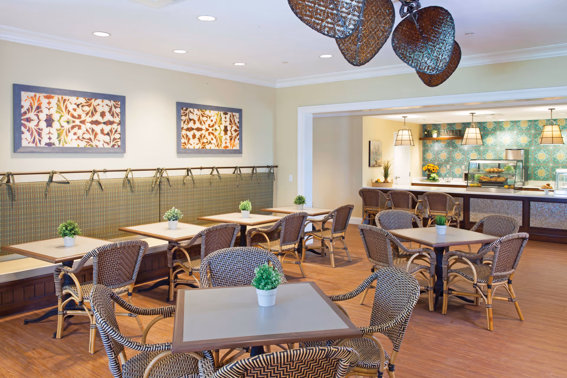 A bright dining area with several square tables and wicker chairs arranged neatly. Each table has a small potted plant as decoration. The back wall features two framed floral artworks above a cushioned bench. In the background, there is a counter area with a display case and hanging pendant lights, decorated with a blue patterned tile backsplash.