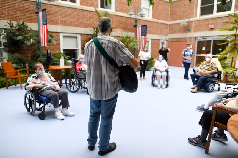 A man playing guitar in an outdoor courtyard area of a senior living facility, surrounded by elderly residents, some in wheelchairs and others seated, all wearing face masks. The courtyard is enclosed by brick walls with windows and American flags hanging on the walls.