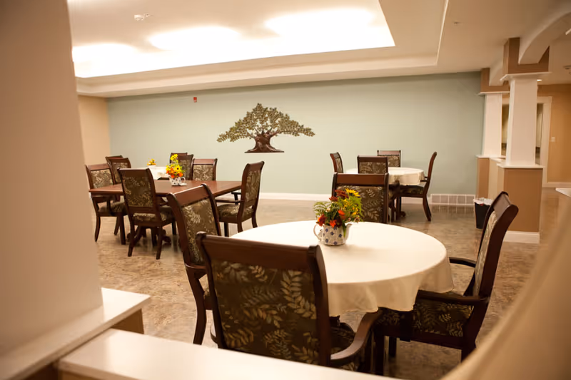 A dining room in Auburn Fields Assisted Living and Memory Care with round and rectangular tables covered with white tablecloths. Each table has a small floral centerpiece. The room has cushioned chairs with patterned upholstery, a light green accent wall with a decorative tree artwork, and soft overhead lighting.