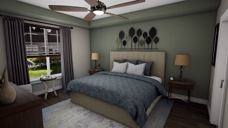 A cozy bedroom with a large bed featuring a beige upholstered headboard and blue patterned bedding. There are two round wooden nightstands on either side of the bed, each with a lamp. A ceiling fan with wooden blades is mounted on the ceiling. The room has a window with dark curtains and blinds, showing a garden outside. A small round table with a chair is placed near the window, and a dresser with decorative vases is visible in the foreground.