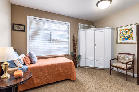 A cozy bedroom with a single bed covered in an orange bedspread and several pillows. Next to the bed is a round wooden side table with a lamp and a small decorative item. A large window with white blinds lets in natural light. Across from the bed is a white wardrobe and a wooden chair with a patterned cushion. The walls are beige, decorated with a framed floral painting and a small sign that reads 'Bless this home'.