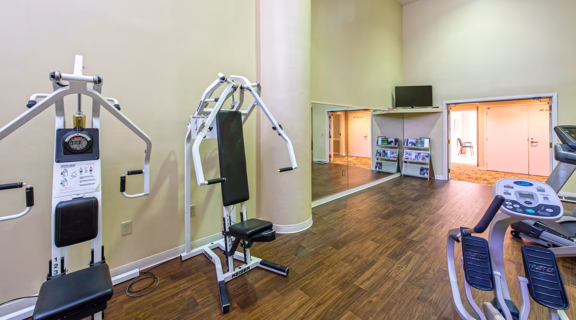 A small fitness room with exercise machines including a chest press, an upper back machine, and a NuStep recumbent cross trainer. The room has wood flooring, beige walls, a large mirror on one wall, a small TV mounted above a shelf, and an open doorway leading to a hallway with carpeted flooring and chairs.