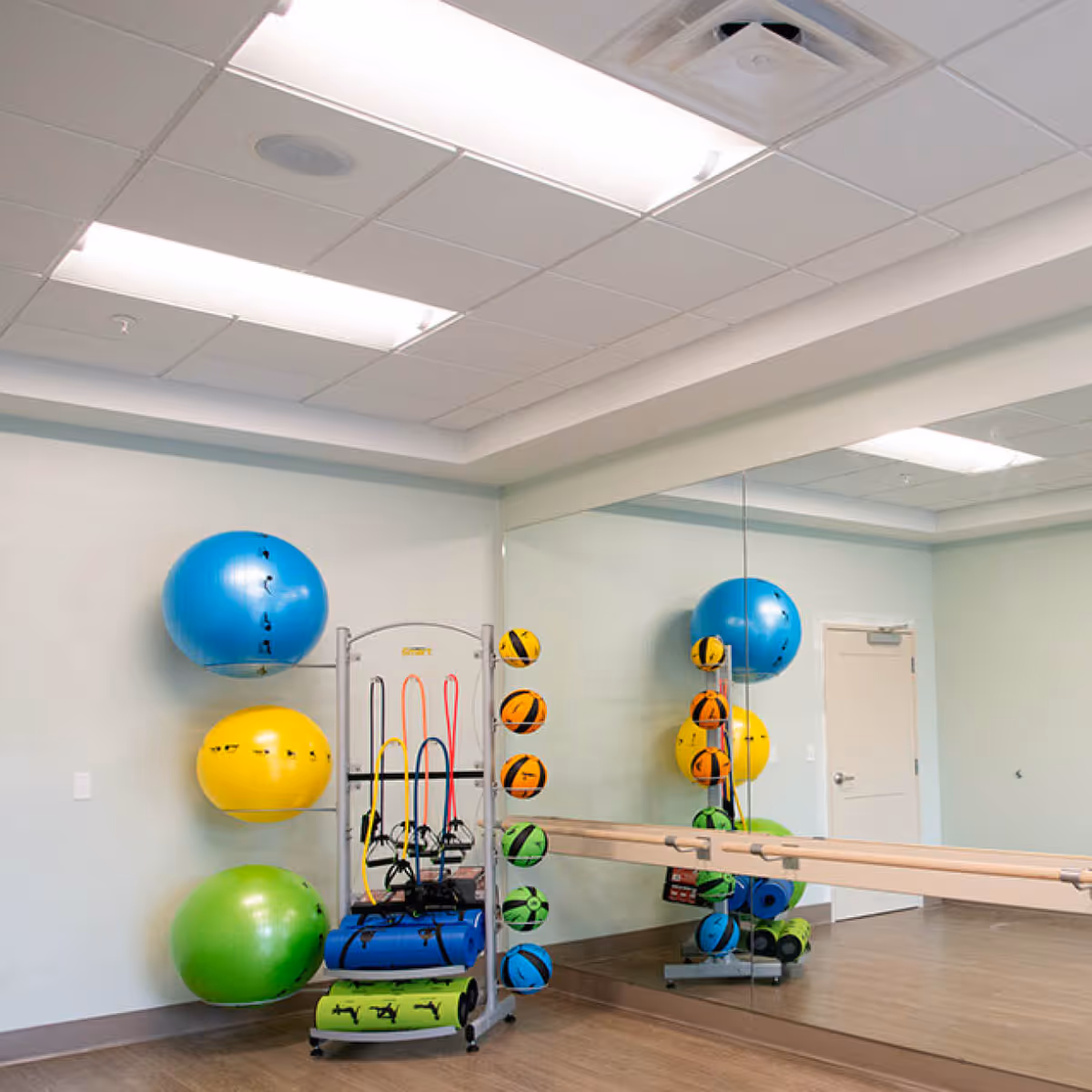 A fitness room with a rack holding colorful exercise balls, resistance bands, and foam rollers. The room has a large mirror on one wall and a wooden ballet barre attached to the mirror. The floor is wooden, and the ceiling has recessed lighting.