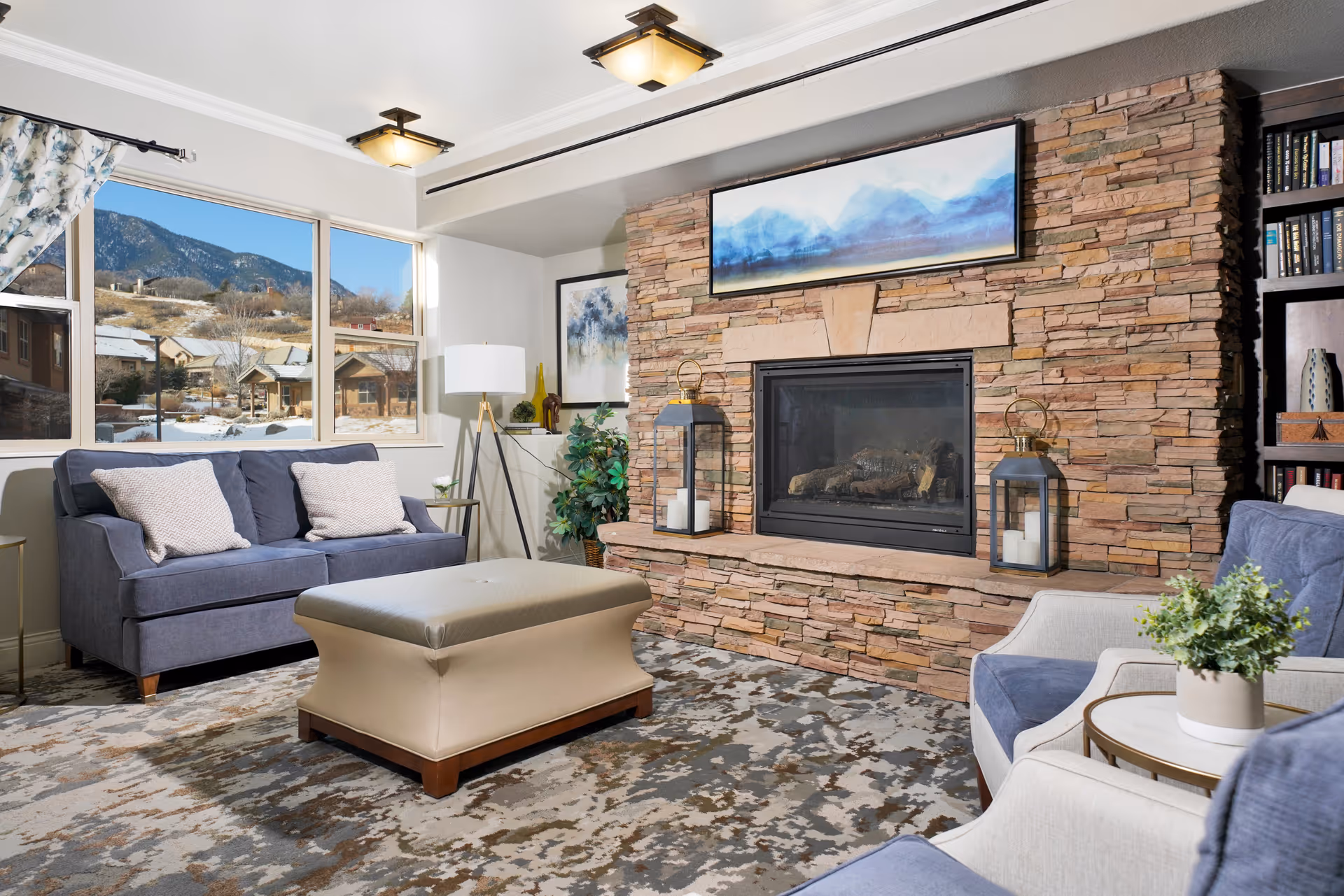 Cozy living room with a stone fireplace, blue sofas, an ottoman, and a window showing snowy mountains.