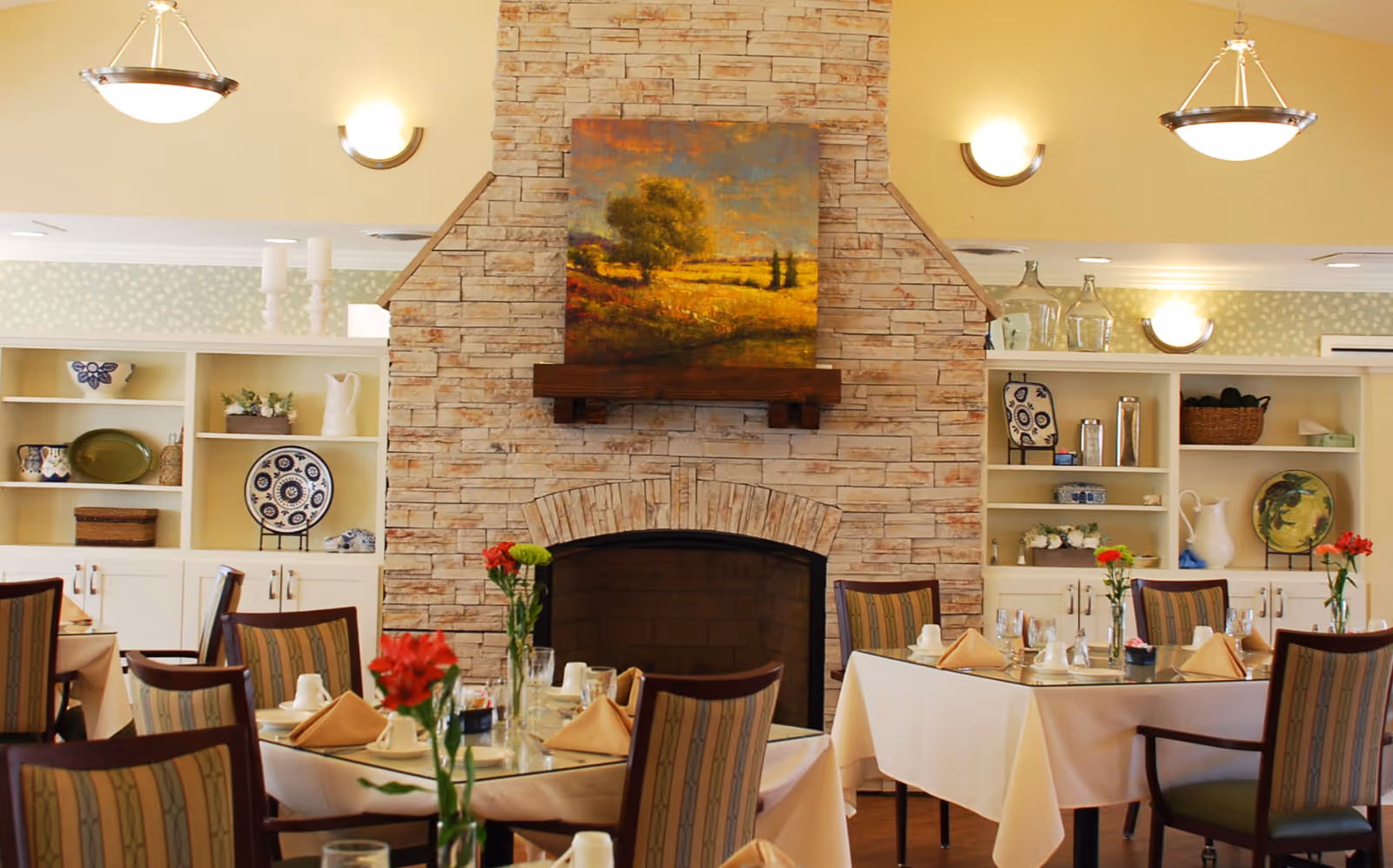 A cozy dining room with tables set for a meal, featuring white tablecloths, folded napkins, glassware, and vases with red flowers. The room has a stone fireplace with a landscape painting above it, built-in shelves with decorative plates and vases, and warm lighting from ceiling fixtures and wall sconces.