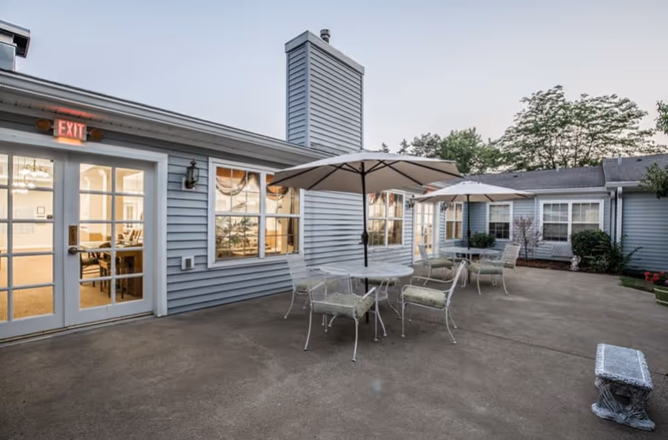 Outdoor patio area at Randall Residence of Tiffin with two round tables, each shaded by a large umbrella, surrounded by cushioned metal chairs. The patio is adjacent to a light blue building with multiple windows and a door with an illuminated exit sign above it. There are some plants and trees visible around the patio.