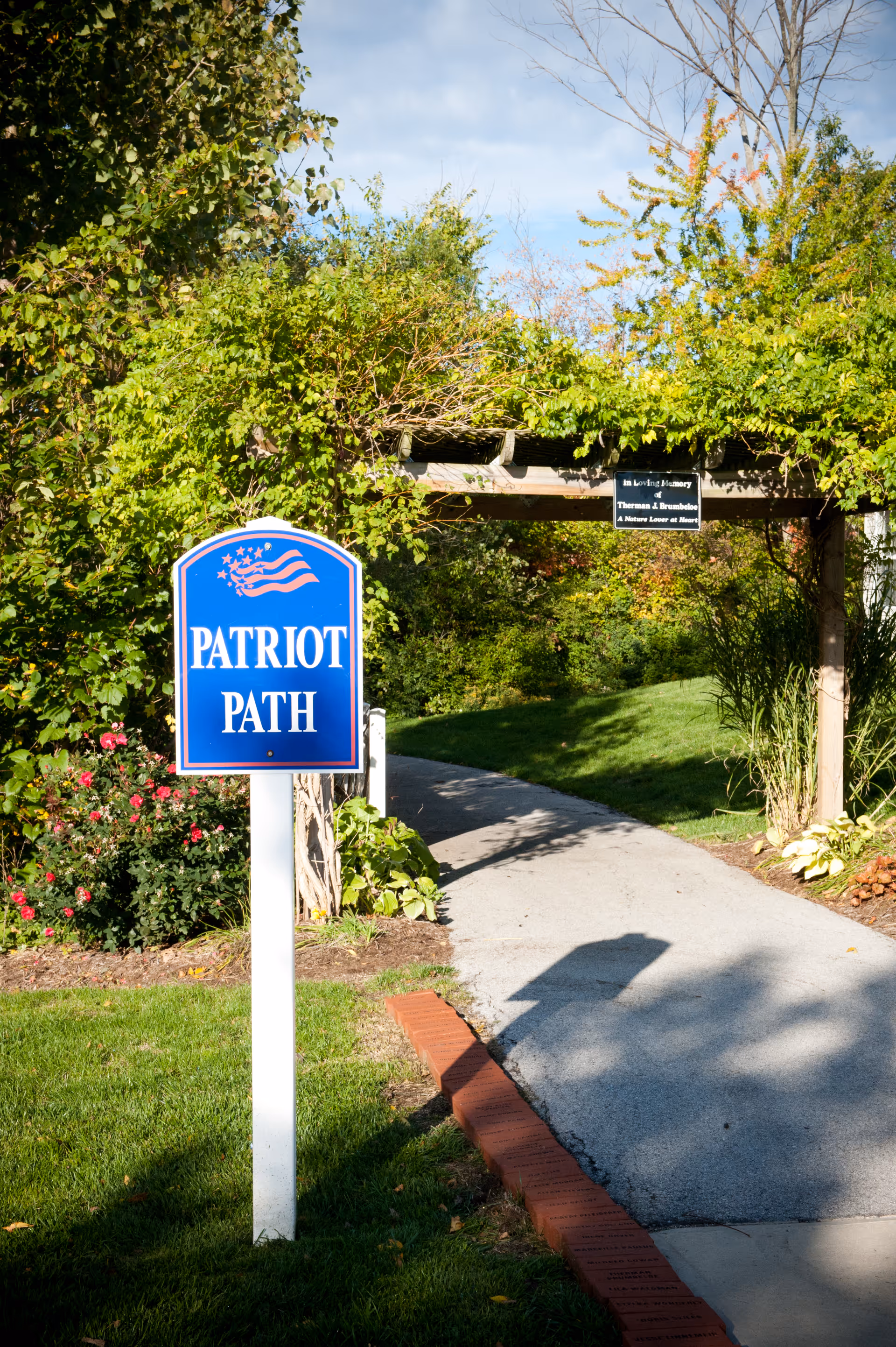 A blue 'Patriot Path' sign marks a paved walkway leading under a leafy arbor in a landscaped garden.