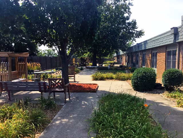 Outdoor patio area at Good Samaritan Society – Ottumwa with concrete walkways, benches, chairs, tables, trees, bushes, and landscaped garden beds alongside a brick building.