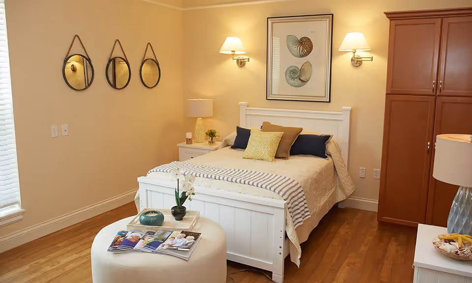 A cozy bedroom with a white wooden bed frame, beige bedding, and multiple pillows in yellow, brown, and navy blue. There are two wall-mounted lamps above the bed and a framed artwork featuring seashells. To the left, three round mirrors hang on the wall. A white ottoman with magazines and a small plant sits at the foot of the bed. The room has wooden flooring and a tall wooden cabinet on the right side.
